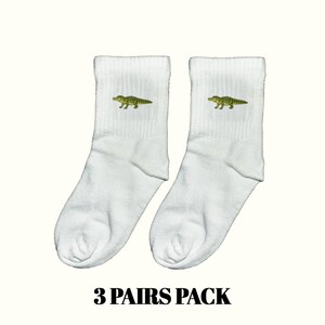 Könnte beinhalten: Ein Paar weiße Socken mit einem kleinen, detaillierten Grafikdruck eines grünen Alligators auf der Außenseite jeder Socke. Der Text 3 PAIRS PACK ist in fetten, schwarzen Großbuchstaben unter den Socken aufgedruckt und gibt die Menge an.