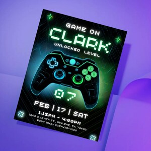 以下が含まれることがあります： 黒色の招待状。ネオンの緑と青のゲームコントローラーのデザイン。テキストは「GAME ON CLARK UNLOCKED LEVEL」と書かれており、イベントの日時と場所が含まれています。招待状は紫色の背景にあります。