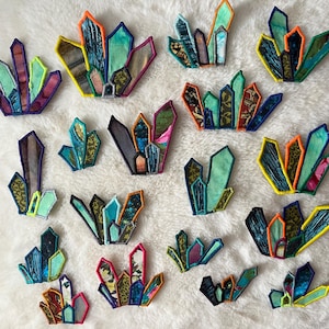 May include: A collection of colorful, embroidered crystal patches. Each patch features a unique design with various shades of blue, green, orange, and purple, outlined with contrasting thread. The patches are arranged on a white, textured surface.