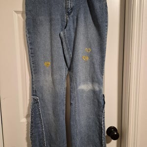 May include: Light blue denim jeans with a frayed waistband and hem. The jeans feature gold heart-shaped embellishments and silver stud detailing along the flared leg openings. A vintage style.