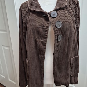 May include: A brown corduroy jacket with a rounded collar and three large, round buttons. The jacket has long sleeves and two front pockets. The fabric has a vertical ribbed texture.