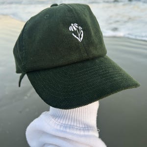 May include: A dark olive green corduroy baseball cap featuring a small, white embroidered flower design on the front centre panel. The cap has a structured crown, a curved brim, and a textured fabric finish with vertical ribbed lines throughout.