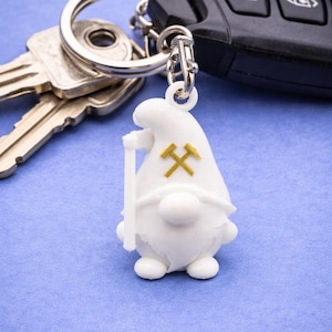 Könnte beinhalten: Ein weißer Zwerg-Schlüsselanhänger mit einem goldenen gekreuzten Spitzhacken- und Hammer-Design. Der Zwerg hält einen weißen Stab. Der Schlüsselanhänger ist an einem silbernen Schlüsselring mit Schlüsseln und einem Autoschlüsselanhänger auf blauem Hintergrund befestigt.