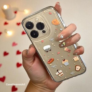 Może przedstawiać: Przezroczyste etui na telefon z różnymi zawieszkami o tematyce jedzenia, w tym chlebem, babeczkami i kawą. Etui ma przezroczysty design z jasnobrązowym tłem i małymi różowymi akcentami w kształcie serca. Widoczny jest tekst "CaseHeavenArt".