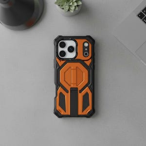 May include: Black and orange protective phone case with a geometric design. The case has a rugged, textured appearance with a combination of black and orange panels. The camera area is visible, and the case appears to be made of durable materials.