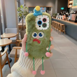 Könnte beinhalten: Eine grüne, flauschige Handyhülle mit einem Cartoon-Gesicht. Die Hülle hat gestickte Augen, einen Mund und einen kleinen blauen Hut mit einem gelben Bommel. Rosa Pompons hängen von unten.