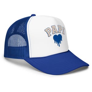 May include: A blue and white trucker cap featuring a white front panel with the word PAPI embroidered in a light beige collegiate font above a blue heart graphic that appears to be dripping. The cap has a blue mesh back and a solid blue brim.