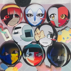May include: A collection of eight circular pin-back buttons featuring stylized anime-inspired character faces with diverse color palettes including red, blue, yellow, and black. Each button displays a unique facial expression and graphic design.