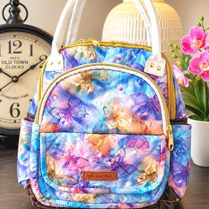May include: A quilted backpack featuring a vibrant blue, purple, and orange butterfly print. It has white top handles, gold-tone zippers, and a brown leather-like patch on the front pocket with the text Hand Made embossed in gold.
