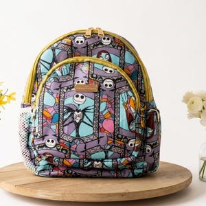 May include: A backpack featuring a repeating pattern of Jack Skellington and Sally from The Nightmare Before Christmas in a stained-glass style design. The bag has gold-toned zippers, a front zippered pocket, and side mesh pockets.