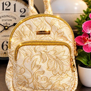May include: A small backpack featuring a cream-colored fabric with a gold floral pattern. The bag includes a top handle, a front zippered pocket, and a gold-toned metal plaque in the center with the word handmade engraved on it.