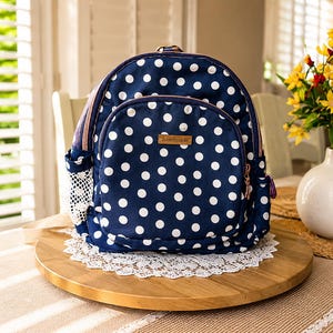 May include: A navy blue backpack featuring a white polka dot pattern, a front zippered pocket, and side mesh pockets. A small rectangular brown label with the word handmade is centered on the front pocket. The bag has rose gold tone zippers.