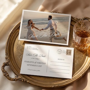 May include: Two white Save The Date cards with elegant script on a gold tray. One card features a photo of a couple on a beach. The other card has the wedding date and names. A glass of amber liquid sits nearby.