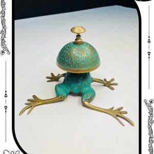 May include: A unique brass frog-shaped bell with a turquoise patina. The bell has a dome-shaped top with intricate patterns and a small knob. The frog's legs are outstretched, giving it a whimsical appearance. The text "RelicForgeCompany" is at the bottom.