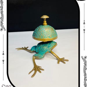 May include: A decorative brass bell with a turquoise patina, shaped like a frog. The bell has an ornate design and a small handle. The frog's legs are outstretched, and the bell sits atop its back. The text "RelicForgeCompany" is at the bottom.