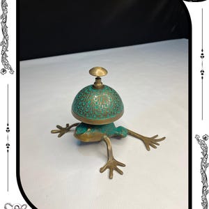 May include: A bronze frog-shaped bell with a turquoise-colored dome-shaped top. The bell has intricate detailing and a small handle. The frog's legs are outstretched, and the bell is resting on a white surface. The text "RelicForgeCompany" is at the bottom.