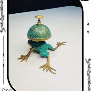 May include: A whimsical frog-shaped bell with a teal and gold finish. The bell has a dome-shaped top with an intricate design. The frog's body and legs are a bronze color. The bell is a decorative item.