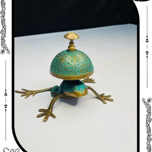 May include: A brass frog-shaped bell with a turquoise and gold dome. The bell has a detailed design and is set on four outstretched legs. The top of the bell has a small brass knob. The base of the image has the text "RelicForgeCompany".