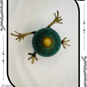May include: A unique, handcrafted bronze frog sculpture. The frog has a teal-colored, patterned body with a central gold-colored accent. It features outstretched bronze legs and the text "RelicForgeCompany" at the bottom.