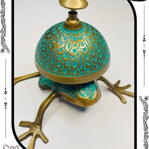 May include: A brass frog-shaped bell with a turquoise and gold dome. The bell has a detailed floral pattern and a small brass knob on top. The frog's legs are extended, and the bell is set against a white background.