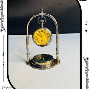 May include: A brass and silver-toned pocket watch with a yellow face and Roman numerals, suspended within a decorative stand. The stand includes a compass at the base. The text "SUPERIOR SHIP TIMEKEEPER" is visible on the watch face. The base of the stand reads "RelicForgeCompany".