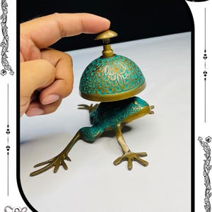 May include: A brass frog-shaped bell with a turquoise patina dome and gold accents. The bell is being pressed by a finger. The base of the bell is a frog with outstretched legs. The words "RelicForgeCompany" are at the bottom.