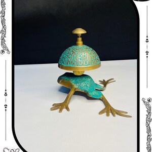 May include: A brass frog-shaped bell with a turquoise and gold dome-shaped top. The bell has intricate detailing and is set against a white and black background. The base of the bell is a frog with outstretched legs.