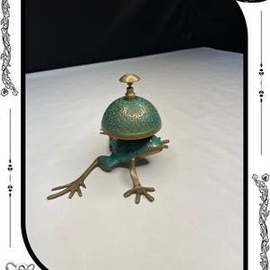 May include: A bronze frog-shaped bell with a verdigris finish. The bell has a dome-shaped top with an ornate design and a small handle. The frog's legs are outstretched, and the bell is resting on a white surface.