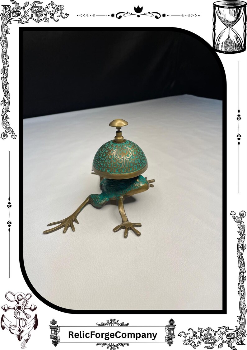 May include: A bronze frog-shaped bell with a verdigris finish. The bell has a dome-shaped top with an ornate design and a small handle. The frog's legs are outstretched, and the bell is resting on a white surface.