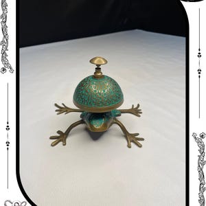 May include: A bronze frog-shaped bell with a turquoise and gold-colored dome. The bell has intricate floral patterns and a small handle on top. The legs are outstretched, giving the bell a whimsical appearance. The text "RelicForgeCompany" is at the bottom.