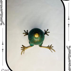 May include: A detailed, antique-style frog sculpture with a teal and gold finish. The frog has a textured body and outstretched legs, with the text "RelicForgeCompany" at the bottom. The sculpture is set against a white background.