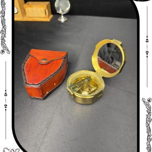 May include: A vintage brass compass with a mirror, open to reveal the compass face. A red leather case with a button closure sits to the left. The compass is a navigational tool, perfect for collectors or those interested in exploration.