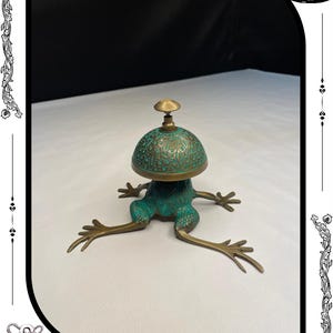 May include: A bronze frog-shaped bell with a green patina and intricate detailing. The bell has a dome-shaped top with a small handle. The frog's legs are outstretched, and the bell is set against a white background.