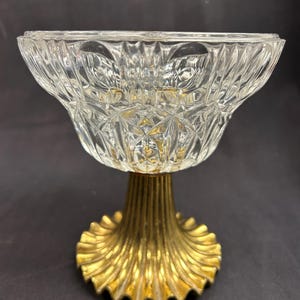 May include: A clear glass bowl with a diamond pattern sits atop a gold-colored, fluted pedestal base. The bowl has a wide, flared shape, and the base has a scalloped edge. The bowl and base are likely vintage.