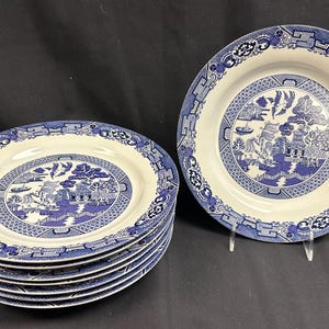 May include: A set of blue and white ceramic plates with a classic Willow pattern. The plates feature a central landscape design and a decorative border. The stack of plates is next to a single plate, both on a black surface.