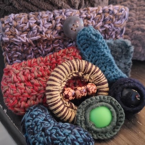 Op de afbeelding: Een verzameling handgemaakte haakartikelen in verschillende kleuren. Bevat een lavendel- en bruine haakpouch met een grijze knoop, een rood haakartikel en een gestreepte bruine en tan haakring met houten kralen. Andere items zijn teal en groen.