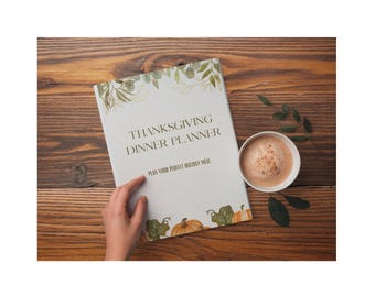 May include: A Thanksgiving Dinner Planner with the text "Thanksgiving Dinner Planner" and "Plan Your Perfect Holiday Meal" is displayed on a wooden surface. The planner has a decorative border of leaves and pumpkins. A cup of coffee sits next to the planner.