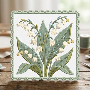 May include: Embroidered lily of the valley design on a square, scalloped-edge fabric. The artwork features white bell-shaped flowers, green leaves, and stems, with a light green border. The background is white, and the design is set on a wooden surface.