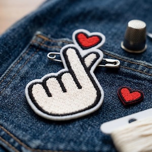 May include: Close-up of a denim pocket with a white and black embroidered hand gesture patch with a red heart. A small red heart patch, thimble, scissors, and thread are also visible.