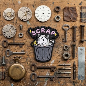 May include: A collection of vintage mechanical parts and ephemera, including clock faces, gears, keys, and a patch with a possum in a trash can that says "SCRAP". The items are arranged on a wooden surface.