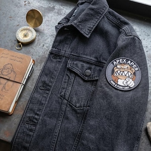 May include: A black denim jacket with a patch that reads "APEX APES". A leather-bound notebook with a gorilla sketch and a compass are next to the jacket. Also present are bullets, a pocket knife, and gloves.