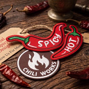 May include: A rustic wooden surface displays red chili peppers and patches. Two red chili pepper patches read "Spicy" and "Hot". A tag reads "Original Spicy Designs". A round patch says "Chili Works".