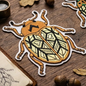 May include: Embroidered patches featuring a stylized beetle design in shades of orange, yellow, and black. The patches are on a wooden surface with a magnifying glass, a sketchbook with a beetle sketch, leaves, and acorns.