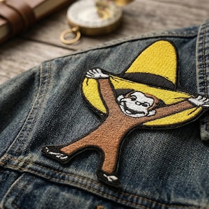 May include: Embroidered patch featuring a cartoon monkey wearing a yellow sombrero, arms outstretched. The patch is brown, tan, black, and white, and is affixed to a denim jacket. The jacket is a medium wash blue.