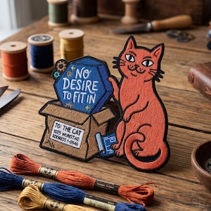 May include: An orange cat patch with the text "NO DESIRE TO FIT IN" next to a cardboard box. The box has the address "TO: THE CAT 1005 WUNSTAD ADDRESS 1-0546". The image also includes spools of thread, scissors, and a leather-bound journal.