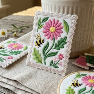 May include: Embroidered floral patches with pink daisies and buzzing bees. The patches have a scalloped edge and are set on a neutral linen surface. A thimble, scissors, and embroidery floss are also visible.