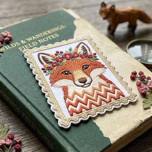 May include: A green book titled "Wilds & Wanderings: Field Notes" with a fox patch. The patch features an embroidered fox wearing a floral crown. Additional elements include a wooden fox figurine, stamps, and a magnifying glass.