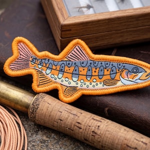 May include: An embroidered patch in the shape of a fish, featuring orange, blue, and pink details. The patch is placed on a leather-bound journal, with a fly fishing rod and a wooden box in the background. The scene evokes a fishing theme.