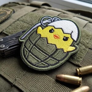 May include: Embroidered patch featuring a yellow chick hatching from a green grenade. The patch is on a green tactical bag, with a multi-tool and several bullets visible. The patch has a white eggshell and a cute chick face.