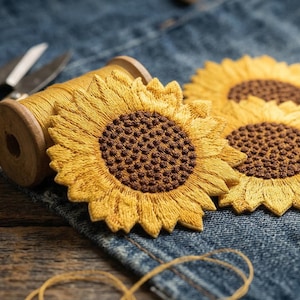 May include: Close-up of embroidered sunflower patches with yellow petals and brown centers. A spool of yellow thread, scissors, and a pin cushion rest on a denim surface, suggesting a crafting theme. The image evokes a sense of handmade artistry.